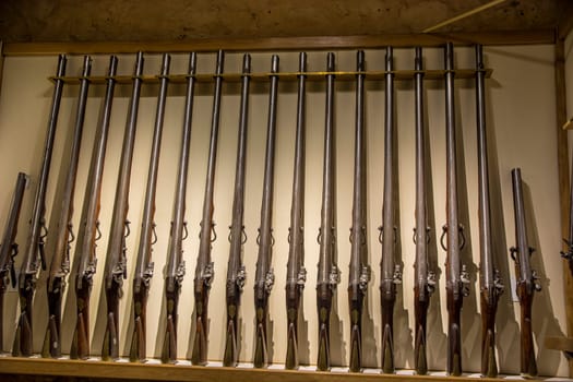 A bunch of guns are lined up on a shelf. Photo of a collection of firearms neatly displayed on a shelf