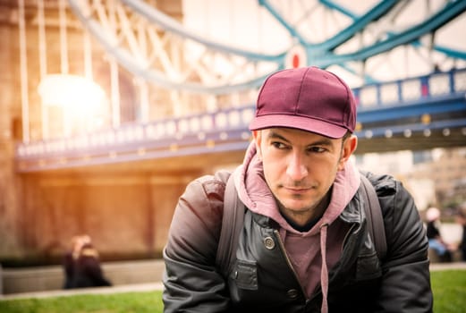 A young man wearing a maroon hat and a black jacket in front of Tower Bridge in London, UK