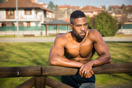 A shirtless muscular african american man leaning on a wooden fence in a city park in a sunny day