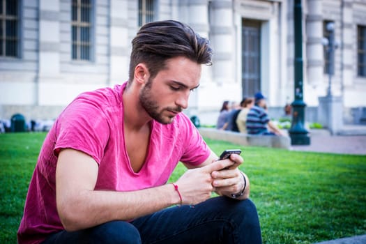 A man sitting on the grass looking at his cell phone. Photo of a man engrossed in his phone while surrounded in a city park