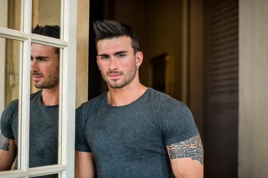 A man standing in front of a window next to another man. Photo of two men standing together by a window, showcasing their muscular and attractive physiques