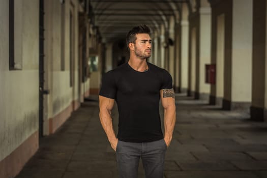 A man in a black shirt standing in a cloister. Photo of a handsome muscular man standing in a colonnade, outdoor in a city, wearing a black shirt