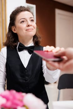 Female concierge eagerly welcomes and assists elderly traveler, checks identification for check-in. At front desk, hotel receptionist receiving passport from retired senior tourist for verification.