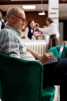 Caucasian senior male client relaxing on comfy sofa with personal computer in hotel lounge area. Retired senior man looking for enjoyable holiday activities on laptop in beautiful resort.
