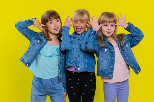 Funny happy teenage girls making playful silly facial expressions and grimacing, fooling around, showing tongue. Little children sisters. Three cute siblings kids isolated on studio yellow background