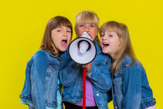 Teenage girls talking with megaphone, proclaiming news, loudly announcing advertisement, warning using loudspeaker to shout speech. Little children sisters. Three siblings kids on yellow background