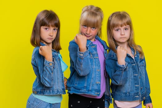 Aggressive angry teenage girls trying to fight at camera, shaking fist, boxing with expression, punishment, abuse, bullying. Little children sisters. Three siblings kids isolated on yellow background