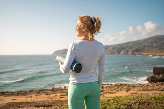 Mature woman with yoga mat standing on coast. Pensioner female looking ocean waves while holding sport mat.Back view fit senior woman wearing sport clothes preparing to do yoga on sea shore