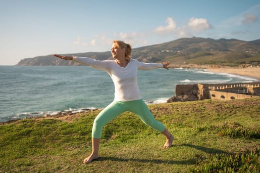 Senior woman practicing yoga on coast. Female standing in warrior position on sea shore with an ancient fortress on background. Mature woman barefoot doing workout near medieval fortification on ocean beach.
