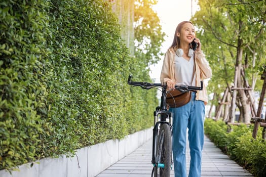 Asian young woman walking in countryside outdoor with her bicycle she using mobile phone talking with friend, Happy female stand on city street with bike using smartphone for summer sightseeing walk