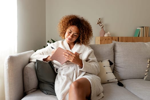 Young multiracial hispanic woman writing on personal journal sitting on the couch at home. African American female writing notes with pen. Lifestyle concept.