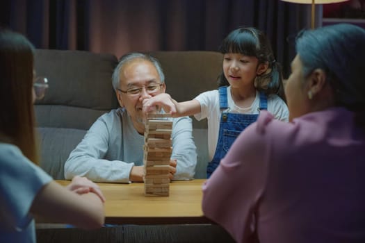 Happy grandparents Asian family enjoy playing toy block with little daughter and mother together at home night time, Smiling parent having fun play build constructor tower of wooden blocks, education