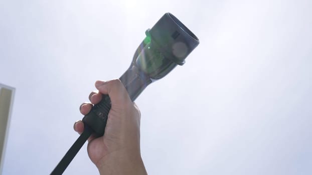 Closeup hand grasping an EV plug for electric vehicle with the midday sky in the background as progressive idea of alternative sustainable clean and green energy for environmental concern.