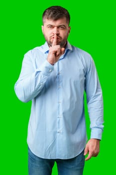 A man in a blue shirt gestures for silence with a finger on his lips against a bright green background in studio