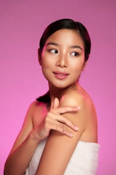 A young woman lightly touches her shoulder while looking back over her shoulder. Soft pink lighting enhances her skin's glow, creating a vibrant atmosphere for the beauty shoot.
