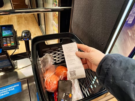 Kirov, Russia - December 03, 2024: Weighing vegetables at a self-service checkout while holding a receipt in a grocery store during evening hours