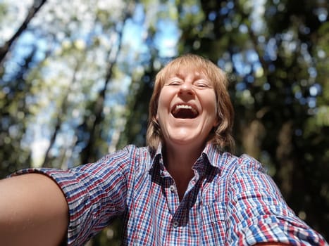 A middle-aged woman with a bright smile takes a joyful selfie among tall trees, experiencing the beauty of nature.