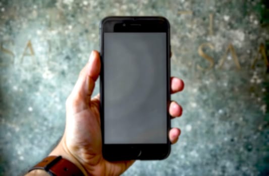 A person presents a smartphone in their hand, showcasing a blank screen against an artistic wall with subtle textures.. Blurred picture