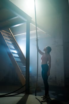A woman stretches her arms in a foggy indoor space reaching Up for gym climbing rope, symbolizing determination and hope for the future