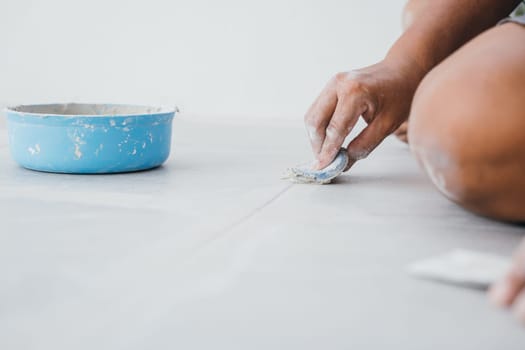 Grouting tiles with a rubber trowel, a hand spreads paste between ceramic tiles on the floor. Tile finishing details in close focus.