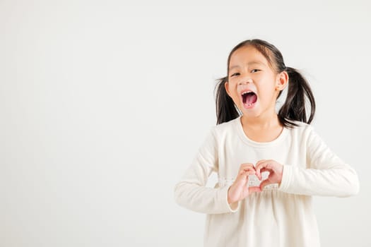 A smiling Asian young kid girl shows confidence, creating a heart shape with fingers and hands on white background. Portrait of kindergarten child sending love for Valentine's Day