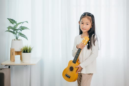 Asian girl strums a ukulele while standing in a cozy, well-decorated living room. Her cheerful expression and love for music harmonize perfectly with the modern, softly lit environment.