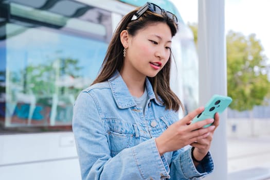 A stylish young woman in a denim jacket checks her smartphone while waiting at a tram station. She appears focused and relaxed, with sunglasses on her head. A modern urban lifestyle scene.