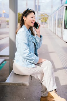 Smiling young woman wearing casual clothes and denim jacket sitting at a tram stop, talking on her mobile phone. Urban setting with tram and city background. Bright and sunny day.