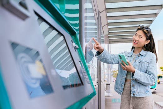 Smiling young woman in a denim jacket using a ticket machine at a tram stop. She holds a smartphone while interacting with the touchscreen. Urban setting with public transport in the background.