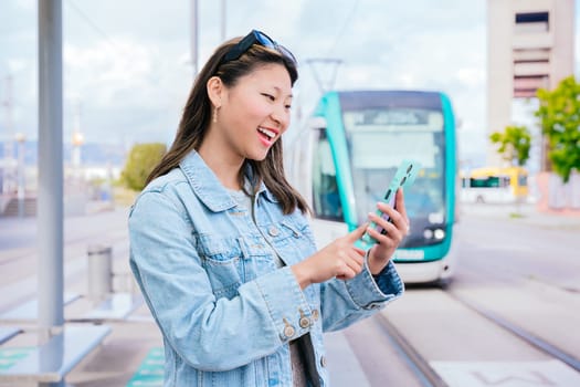 Cheerful young woman wearing a denim jacket using her smartphone while waiting at a tram stop. A modern tram is visible in the background. Bright and lively urban environment.