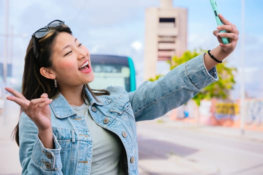 Cheerful young woman in a denim jacket taking a selfie with her smartphone at a tram stop. She smiles and makes a peace sign, with a tram and urban background behind her.