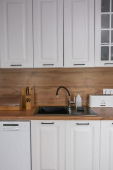 Horizontal photo of a modern kitchen in white and wooden style, in the center is a black kitchen sink, on a wooden countertop there are white wooden appliances for storing and preparing food. Modern kitchen concept, kitchen island, modern kitchen style. High quality photo