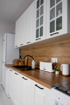 Horizontal photo, side view of a modern kitchen with a white cabinet and a wooden countertop on which there is a toaster, plants and a bread box with a knife rack, a prominent gas stove, a black sink and a white refrigerator. Modern kitchen concept, kitchen island, modern kitchen style. High quality photo