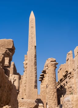 Ancient Egyptian obelisk covered in hieroglyphs stands tall against a clear blue sky, surrounded by ruins of Karnak temple