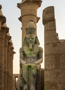 A massive stone statue of Ramesses II stands in Luxor temple, showcasing intricate carvings under the warm glow of the setting sun.