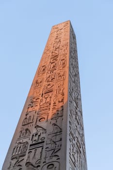 Ancient Egyptian obelisk standing tall against a clear blue sky, its surface covered in intricate hieroglyphs. Placed at the entrance of Luxor temple
