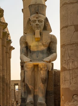 A massive stone statue of Ramesses II stands in Luxor temple, showcasing intricate carvings under the warm glow of the setting sun.