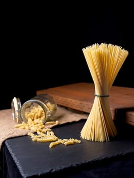 A still life photo of dried spaghetti noodles standing up and spiral noodles spilling out of a glass jar.