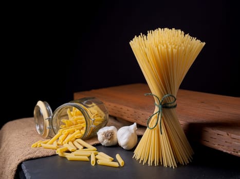 A still life photograph of dried penne pasta in a glass jar, dried spaghetti noodles in a bundle and garlic bulbs.