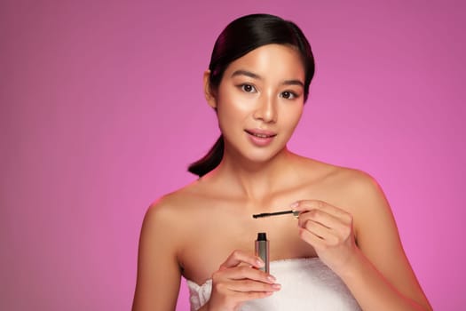 A model holds a mascara brush, preparing to apply makeup while showcasing her natural beauty. The vibrant pink backdrop enhances the focus on her skincare routine.