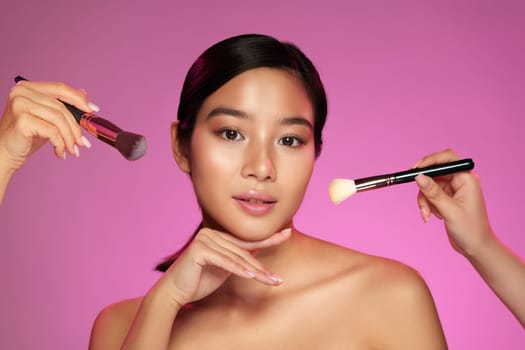 A model poses with a serene expression as makeup artists apply products using brushes. The bright pink background enhances the beauty-focused atmosphere and highlights the artistry involved.