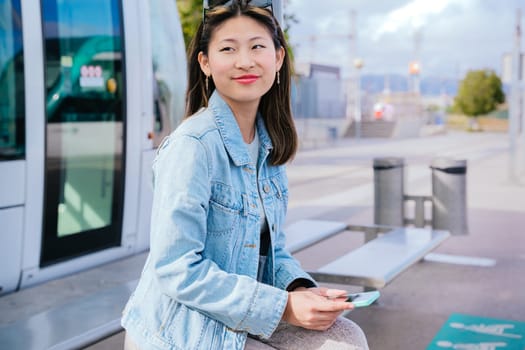 Stylish young woman in a denim jacket sitting at a tram stop, holding her smartphone. She looks to the side with a relaxed expression. Modern urban transportation and city life concept.