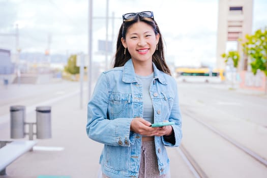 Cheerful young woman in a denim jacket holding her smartphone while looking up with a happy expression. Modern urban setting, representing digital connectivity and city lifestyle.