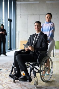 In a professional office setting, a businesswoman helping her director, who is sitting in a wheelchair and using a tablet, while their colleagues collaborate in the background