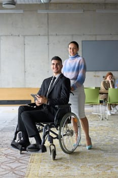 In a professional office setting, a businesswoman helping her director, who is sitting in a wheelchair and using a tablet, while their colleagues collaborate in the background