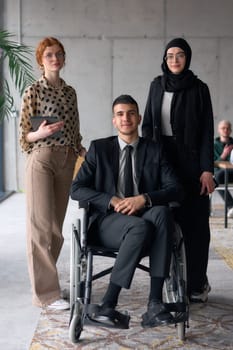 A diverse business team poses confidently, including a smiling director seated in a wheelchair, flanked by two professional female colleagues, one wearing a hijab and the other holding a tablet, in a modern office environment.