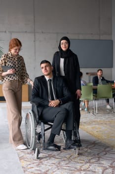 A diverse business team poses confidently, including a smiling director seated in a wheelchair, flanked by two professional female colleagues, one wearing a hijab and the other holding a tablet, in a modern office environment.