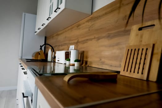 View of the wooden countertop in a modern kitchen in white and wood tones, on which there are wooden cutting boards and coasters, plants, a kettle and toaster, a black sink, a wooden knife holder and a bread box. Concept of modern functional kitchen, kitchen design, convenience and comfort. High quality photo