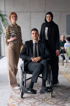 A diverse business team poses confidently, including a smiling director seated in a wheelchair, flanked by two professional female colleagues, one wearing a hijab and the other holding a tablet, in a modern office environment.