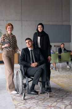 A diverse business team poses confidently, including a smiling director seated in a wheelchair, flanked by two professional female colleagues, one wearing a hijab and the other holding a tablet, in a modern office environment.